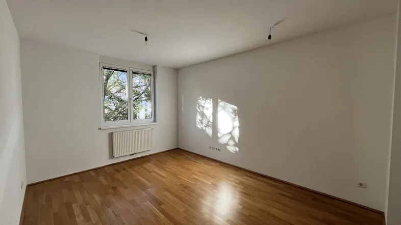 Helles Zimmer mit Parkettboden und einem Fenster, das viel Tageslicht hereinlässt.