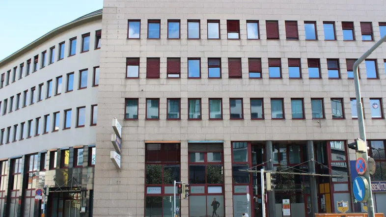 Modernes Bürogebäude mit mehreren Etagen, großen Fenstern und roten Fensterläden an einer Straße.