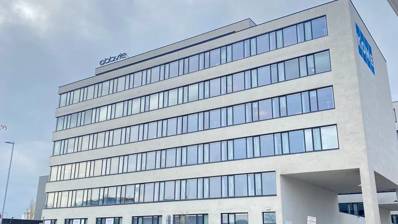 Modernes Bürogebäude mit vielen Fenstern und klaren Linien unter bewölktem Himmel, umgeben von einer städtischen Umgebung.