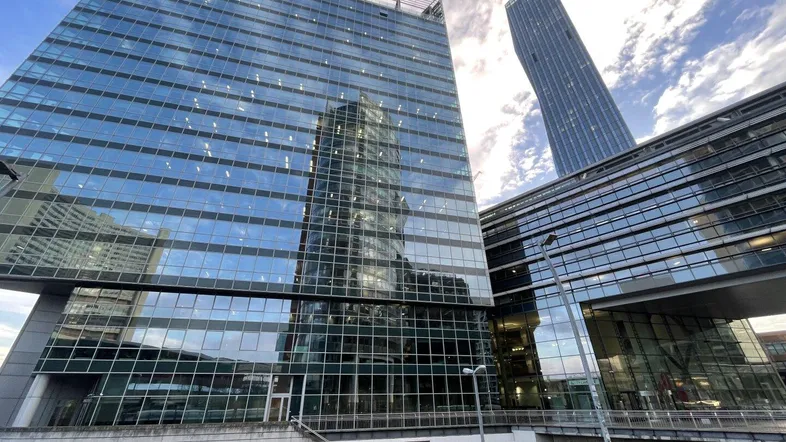Moderne Glasfassade eines Bürogebäudes mit einem weiteren Wolkenkratzer im Hintergrund unter bewölktem Himmel.