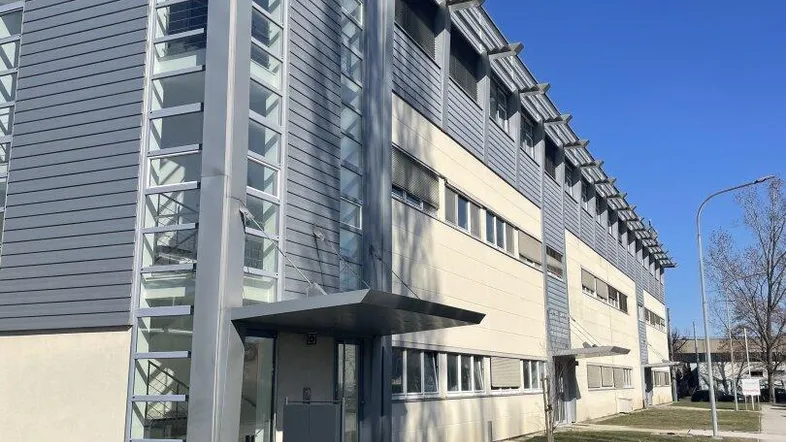 Modernes Bürogebäude mit Glasfassade und Metallverkleidung unter blauem Himmel, umgeben von Grünflächen.