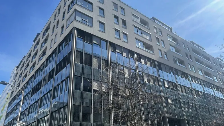 Modernes Bürogebäude mit großer Glasfassade und mehreren Etagen unter blauem Himmel, das eine städtische Umgebung zeigt.