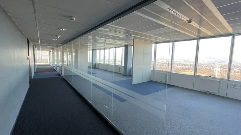 Langer Bürogang mit Glaswänden und Teppichboden, der zu einem offenen Bürobereich mit Stadtblick führt.