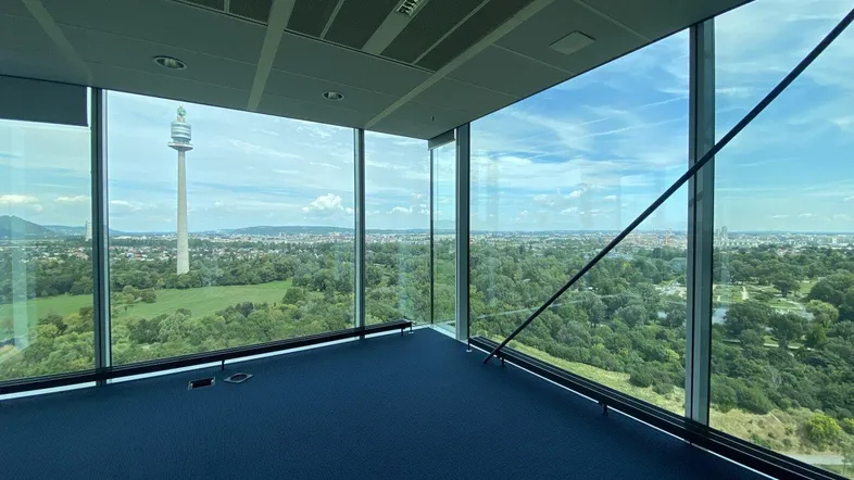 Geräumiges Büro mit blauer Teppichboden und raumhohen Fenstern, die einen weiten Blick über die Stadt bieten.