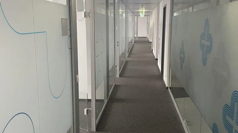 Langer Bürogang mit Glaswänden, der zu mehreren Büroräumen führt und eine moderne Arbeitsumgebung schafft.