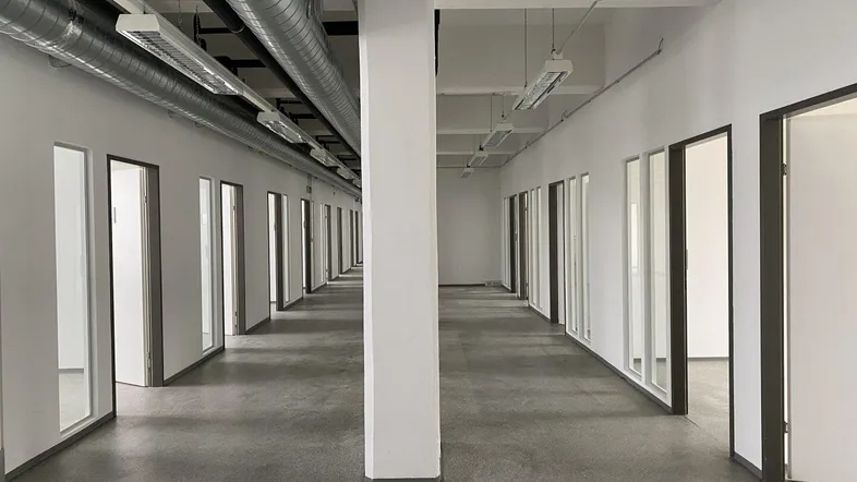 Heller Bürogang mit einer Reihe von Bürotüren und einem zentralen Pfeiler, gut beleuchtet und mit sichtbaren Rohren.