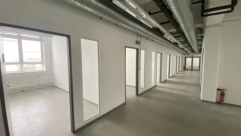 Langer Bürogang mit vielen einzelnen Büroräumen, ausgestattet mit großen Fenstern und sichtbaren Lüftungsrohren an der Decke.