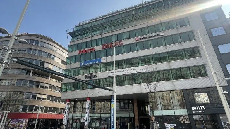 Moderne Bürogebäude-Fassade mit großen Glasflächen und Geschäften im Erdgeschoss in einer belebten Stadtumgebung.