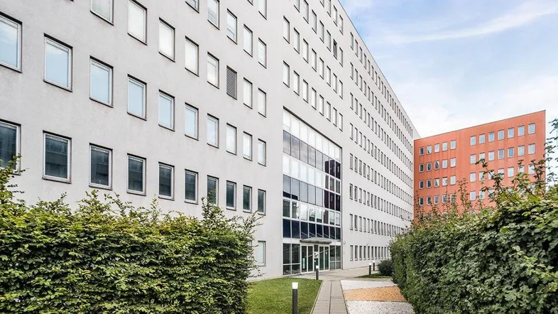 Moderne Bürogebäude-Fassade mit vielen Fenstern, umgeben von gepflegten Hecken und einem Gehweg.