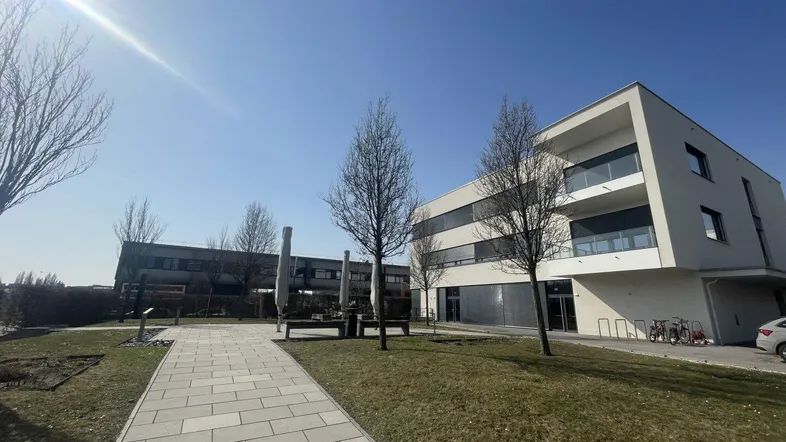 Modernes Gebäude mit klarer Architektur, umgeben von Grünflächen und einem gepflasterten Weg unter blauem Himmel.