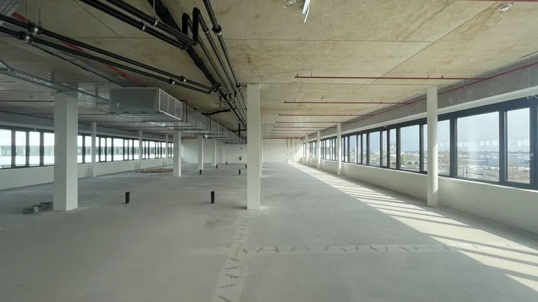 Großer, leerer Büroraum mit Betonboden und Decke, sichtbaren Leitungen und vielen Fenstern.