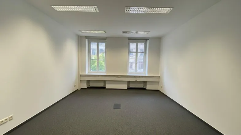Leeres Büro mit Teppichboden, weißen Wänden und zwei Fenstern mit Jalousien, die für Helligkeit sorgen.