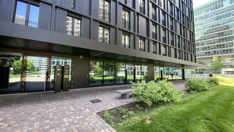 Eingangsbereich des modernen Gebäudes mit großen Glasfronten, gepflastertem Vorplatz und Grünanlagen.