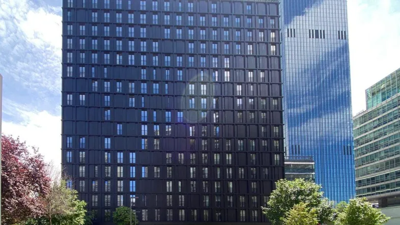 Modernes, dunkles Hochhaus mit vielen Fenstern und einer gepflegten Grünanlage davor, unter blauem Himmel.