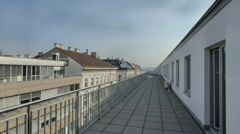 Lange, schmale Dachterrasse mit Fliesenboden und Metallgeländer, umgeben von städtischer Architektur und Gebäuden.