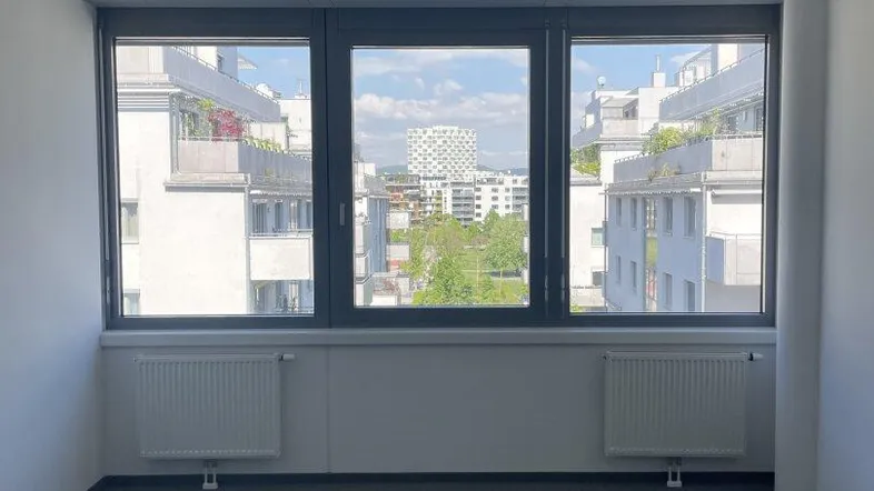 Geräumiges, leeres Büro mit großen Fenstern, die einen Blick auf umliegende Gebäude und Grünflächen bieten.