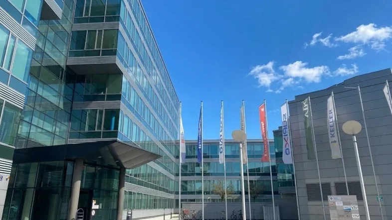 Modernes Bürogebäude mit Glasfassade und Firmenflaggen unter blauem Himmel, das eine professionelle Atmosphäre ausstrahlt.