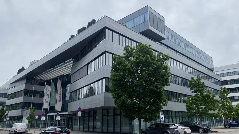 Modernes Bürogebäude mit Glasfassade und mehreren Etagen, umgeben von Bäumen und Parkplätzen.