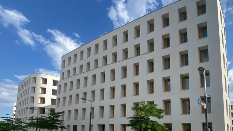 Modernes weißes Gebäude mit vielen Fenstern, umgeben von Bäumen und einer Straße mit parkenden Autos.