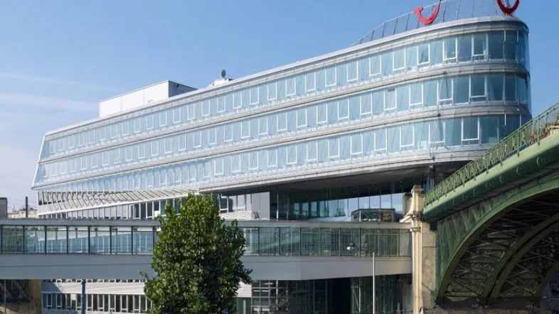 Modernes Bürogebäude mit geschwungener Glasfassade und angrenzender Brückenkonstruktion unter blauem Himmel.