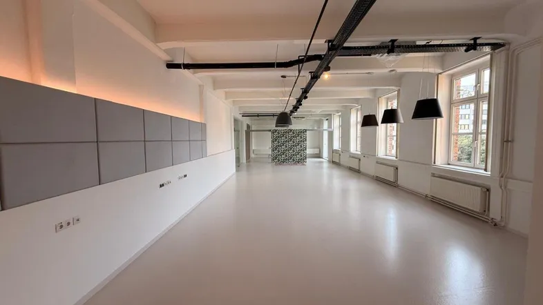 Geräumiger, heller Büroraum mit hohen Decken, großen Fenstern und moderner Beleuchtung.