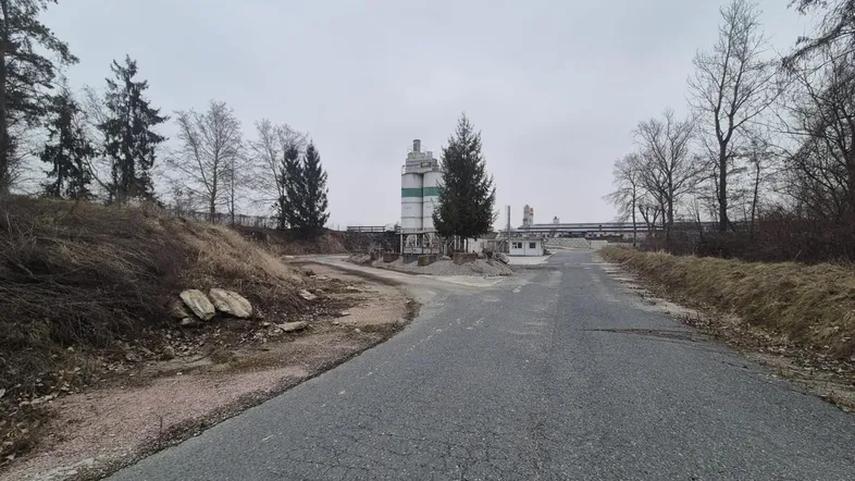 Eine Zufahrtsstraße führt zu einem Industriegelände mit Silos und umliegender Vegetation unter bewölktem Himmel.