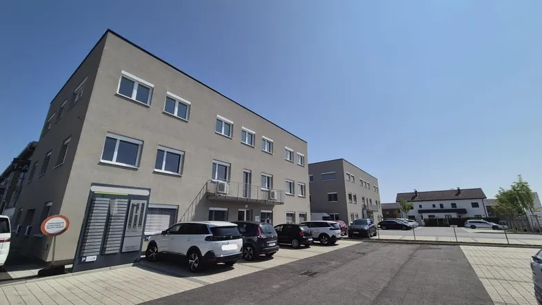 Modernes Geschäftsgebäude mit grauer Fassade und zahlreichen Parkplätzen unter blauem Himmel.