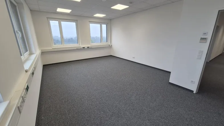 Geräumiges, leeres Büro mit grauem Teppichboden, weißen Wänden und mehreren Fenstern für viel Tageslicht.