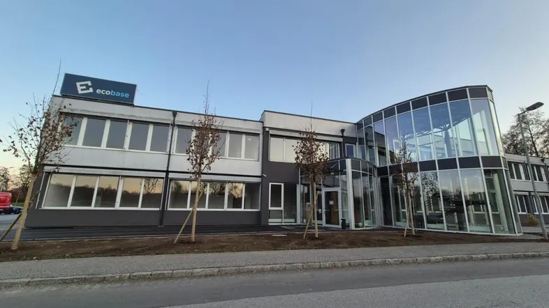 Modernes Bürogebäude mit Glasfassade und "ecobase"-Beschriftung, umgeben von Bäumen und einer Straße.