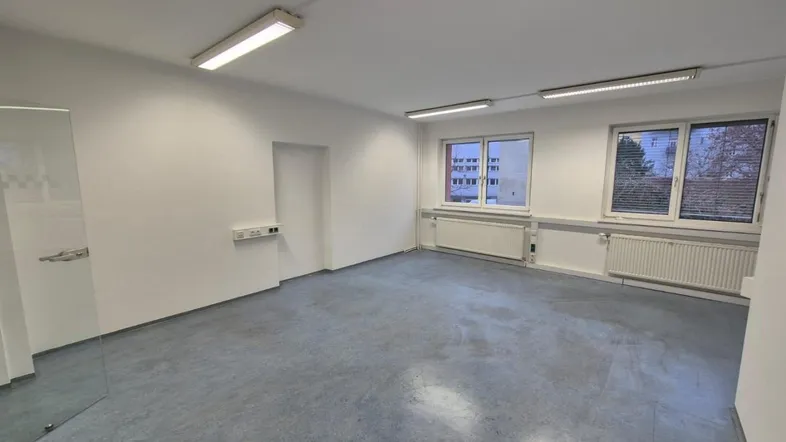 Geräumiges, leeres Büro mit großen Fenstern, grauem Boden und modernen Deckenleuchten.
