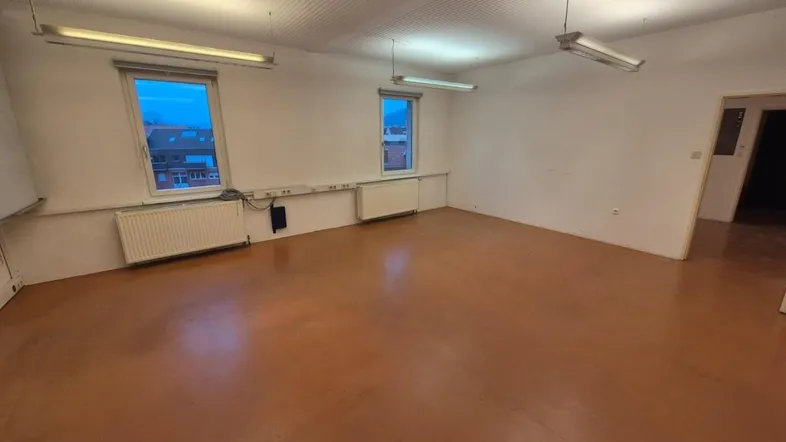 Geräumiger, leerer Raum mit zwei Fenstern, Heizkörpern und einfacher Deckenbeleuchtung, ideal als Büro oder Lager.