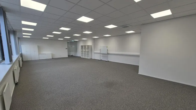 Geräumiges, leeres Büro mit Teppichboden, großen Fenstern und abgehängter Decke mit integrierten Leuchten.