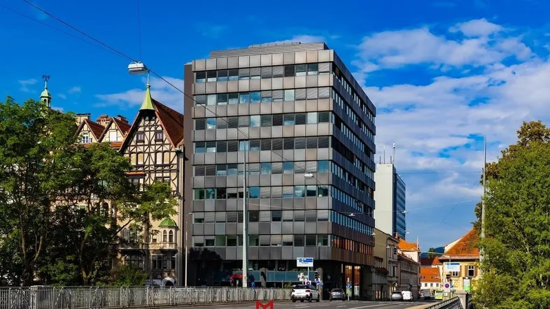 Modernes Bürogebäude mit Glasfassade an einer Brücke, umgeben von älteren Gebäuden und Bäumen unter blauem Himmel.