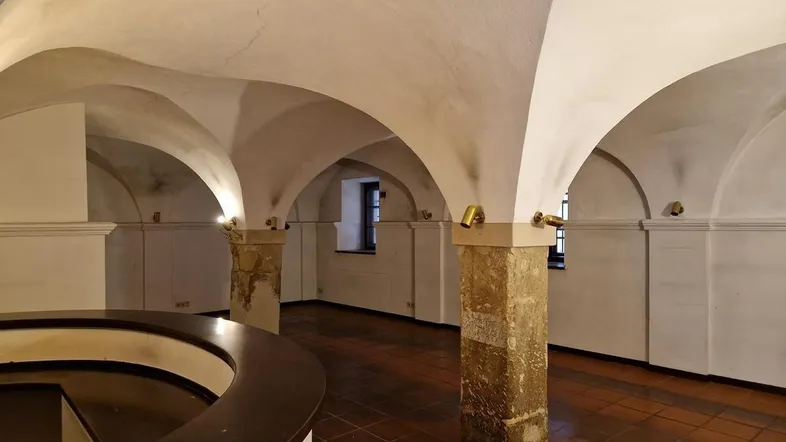 Historischer Gewölbekeller mit robusten Steinsäulen und Terrakottafliesen, beleuchtet durch kleine Fenster.