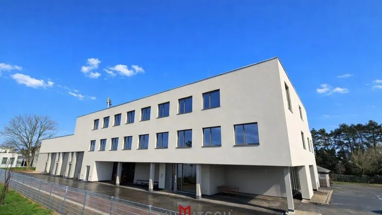 Modernes Gebäude mit heller Fassade, vielen Fenstern und überdachtem Eingangsbereich unter blauem Himmel.