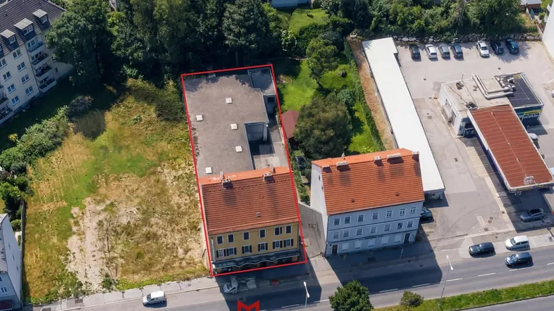 Luftaufnahme eines Gebäudekomplexes mit roter Umrandung in städtischer Umgebung, umgeben von Grünflächen und Parkplätzen.