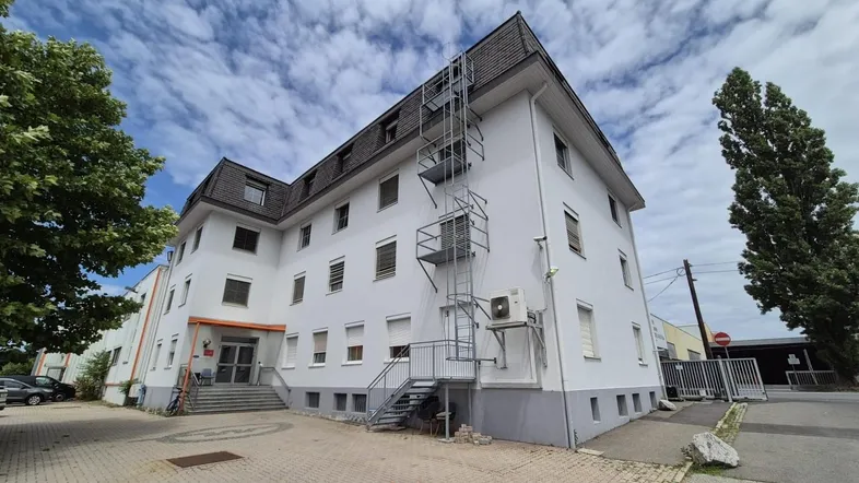 Mehrstöckiges Gebäude mit weißer Fassade, dunklem Dach und externer Metalltreppe unter bewölktem Himmel.