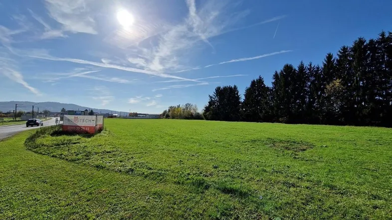 Weitläufiges grünes Grundstück unter blauem Himmel mit Fernblick auf Berge und Bäume.