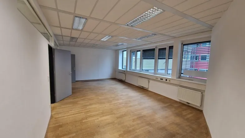 Geräumiges Büro mit Holzparkettboden, weißen Wänden und großen Fenstern mit Jalousien.