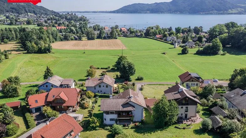 Luftaufnahme einer Wohnsiedlung mit Häusern, grünen Feldern und einem See mit Bergen im Hintergrund.