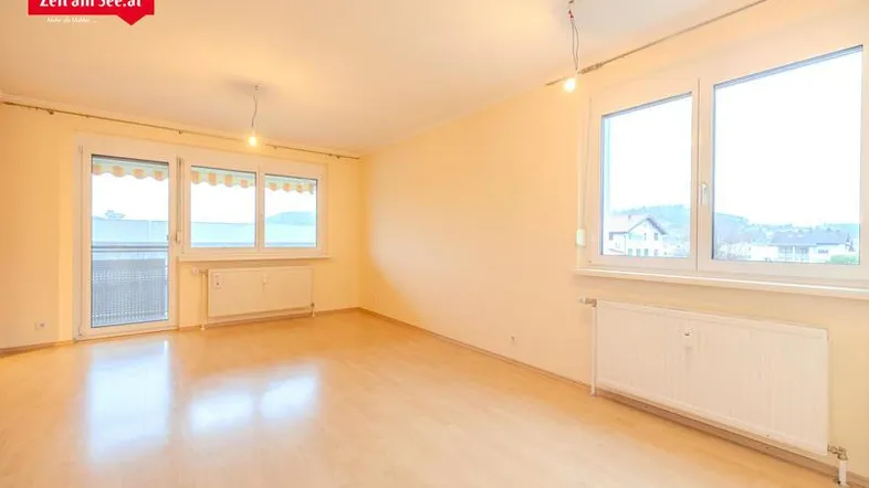 Helles, leeres Wohnzimmer mit Laminatboden, großen Fenstern und Zugang zum Balkon.