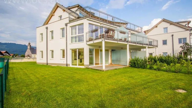 Modernes Wohngebäude mit großem Garten, Holzterrasse und Balkonen unter blauem Himmel.