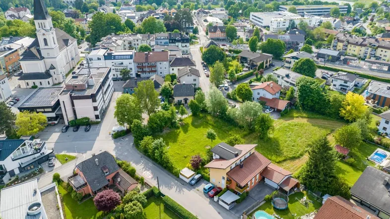 Weitläufige Luftaufnahme einer grünen Wohngegend mit Einfamilienhäusern, Gärten und einer markanten Kirche im Zentrum.