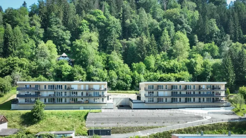 Zwei moderne Apartmentgebäude mit Balkonen, eingebettet in eine grüne Hügellandschaft mit Waldblick.