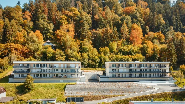 Zwei moderne Apartmentgebäude eingebettet in eine herbstliche Waldlandschaft mit bunten Bäumen.