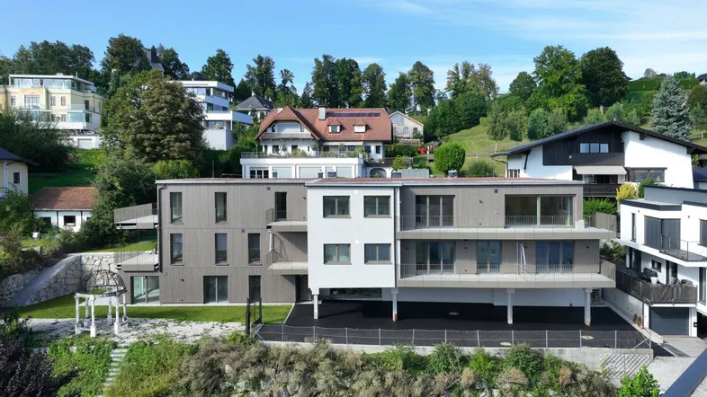 Modernes Mehrfamilienhaus mit Balkonen und Terrassen in grüner Hanglage unter blauem Himmel.