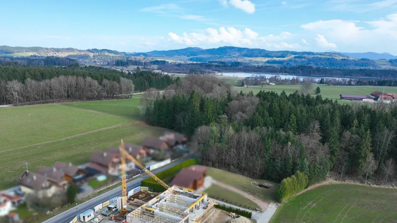 Luftaufnahme einer Neubau-Baustelle mit weitem Blick über Felder, Wälder, einen See und Berge.