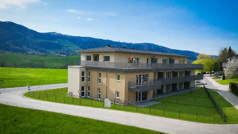 Modernes Mehrfamilienhaus mit Balkonen, umgeben von grünen Wiesen und Bergblick unter blauem Himmel.