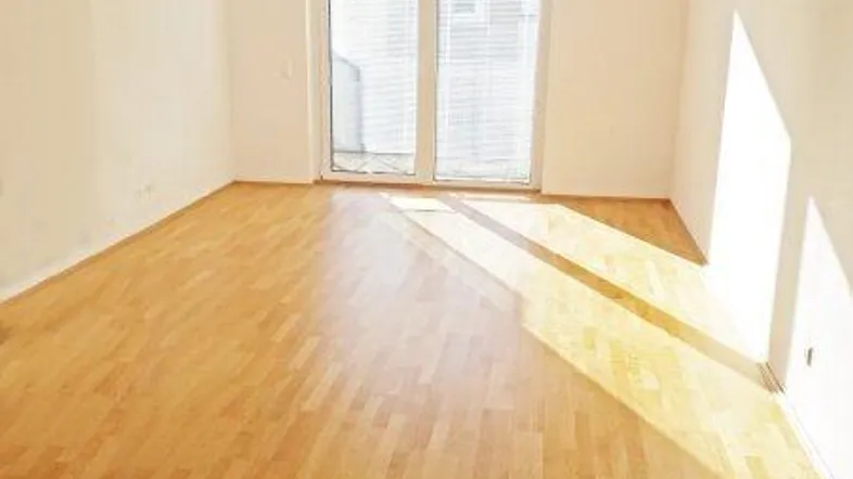 Helles, leeres Zimmer mit Holzboden und Zugang zu einem Balkon oder einer Terrasse.