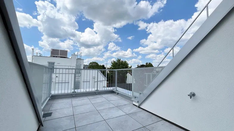 Weitläufige Dachterrasse mit grauen Fliesen und robustem Metallgeländer unter einem blauen Himmel mit Wolken.